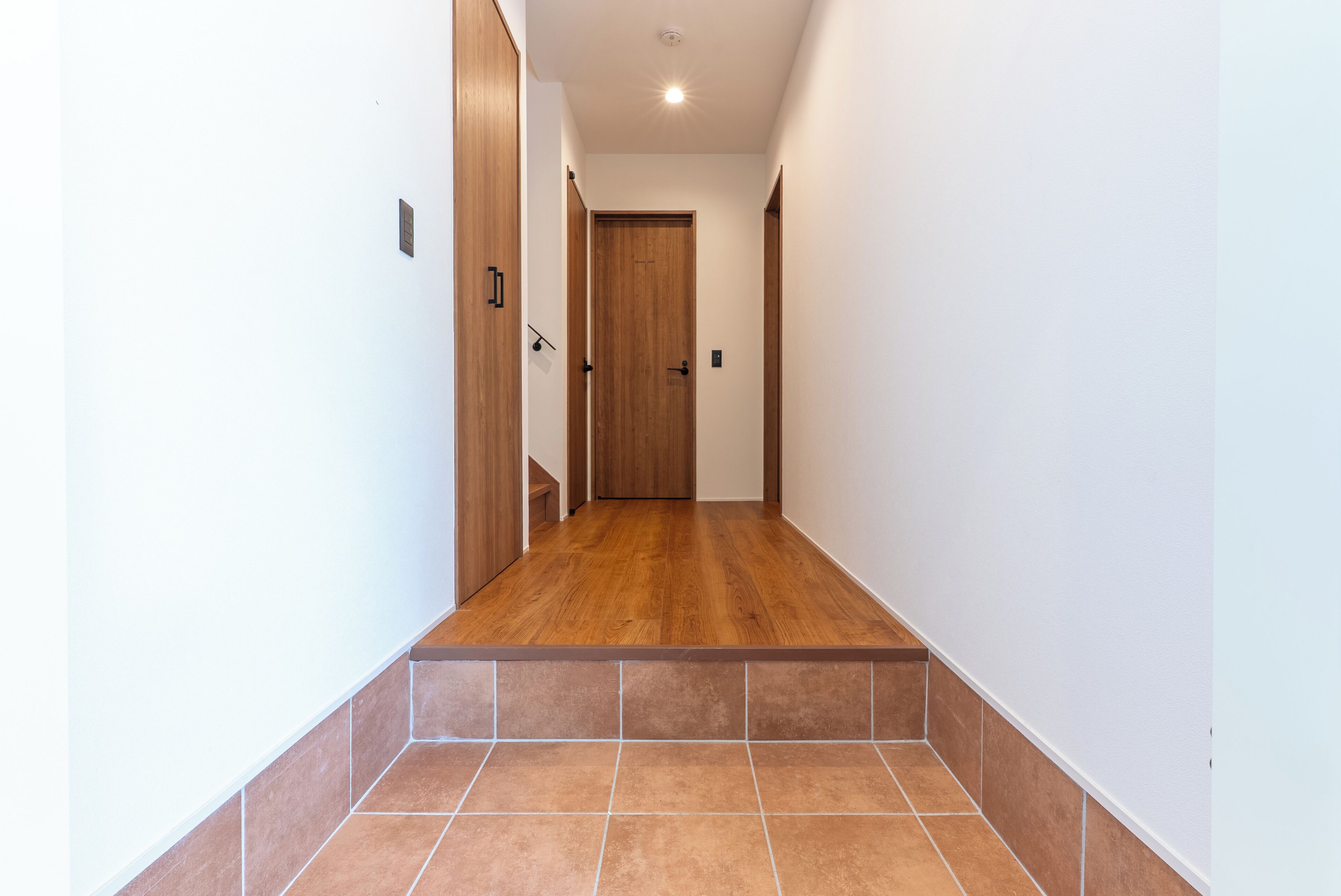Un pasillo con una puerta de madera al final, suelo de baldosas y un accesorio de iluminación en el techo.