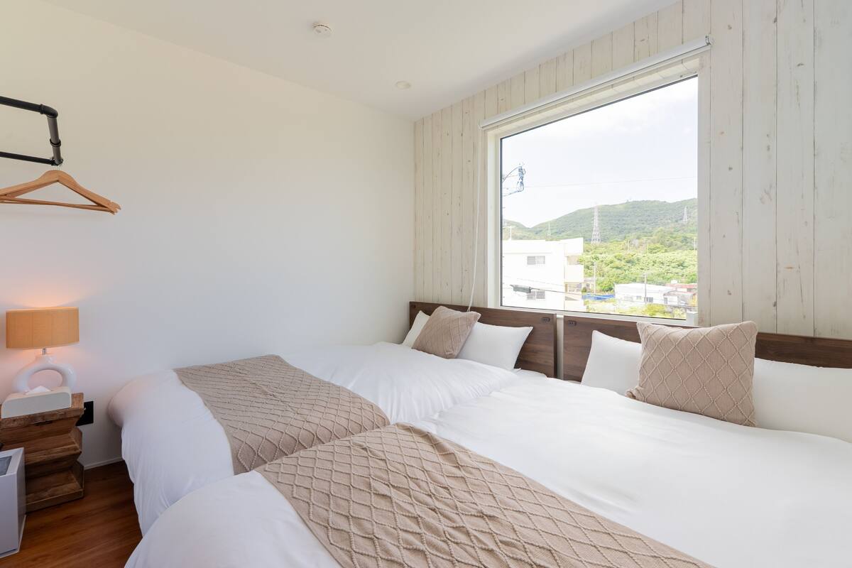 A hotel room with two beds, a wooden headboard, a large window, and a bedside table with a lamp.