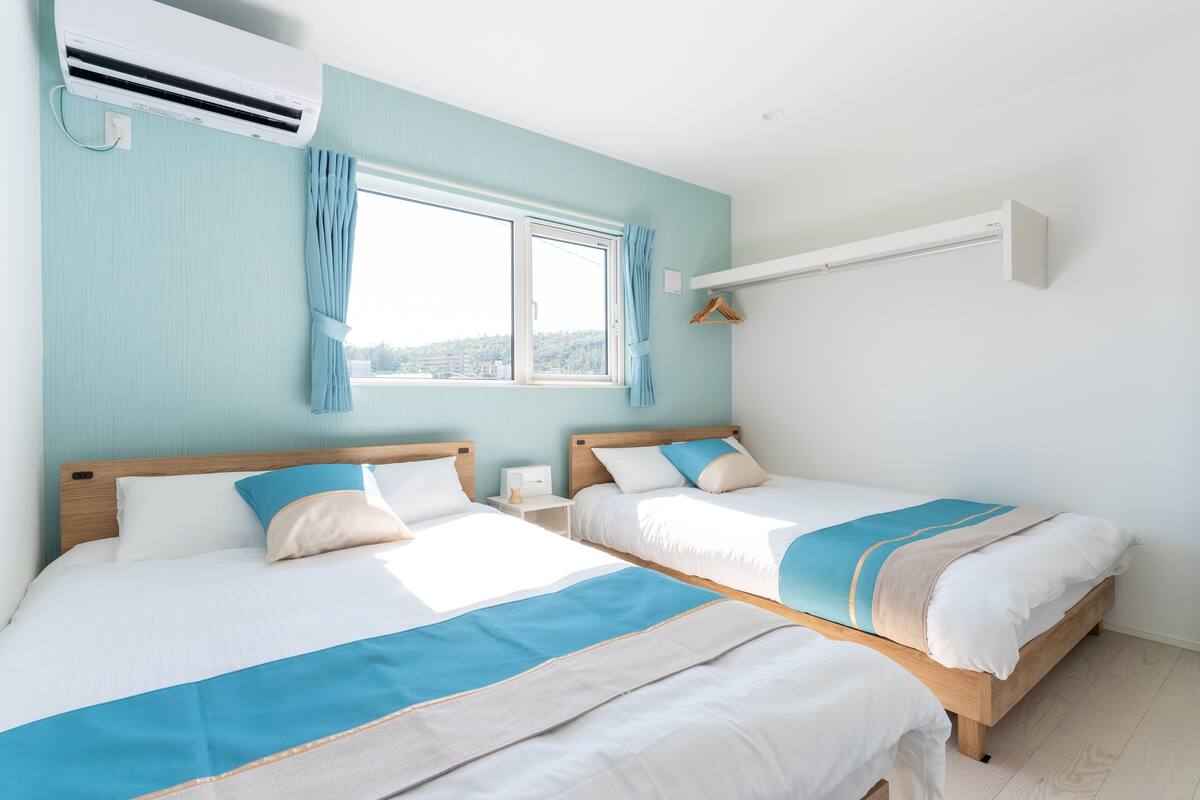 A room with two beds, a window with curtains, an air conditioning unit, and a hanging coat rack.
