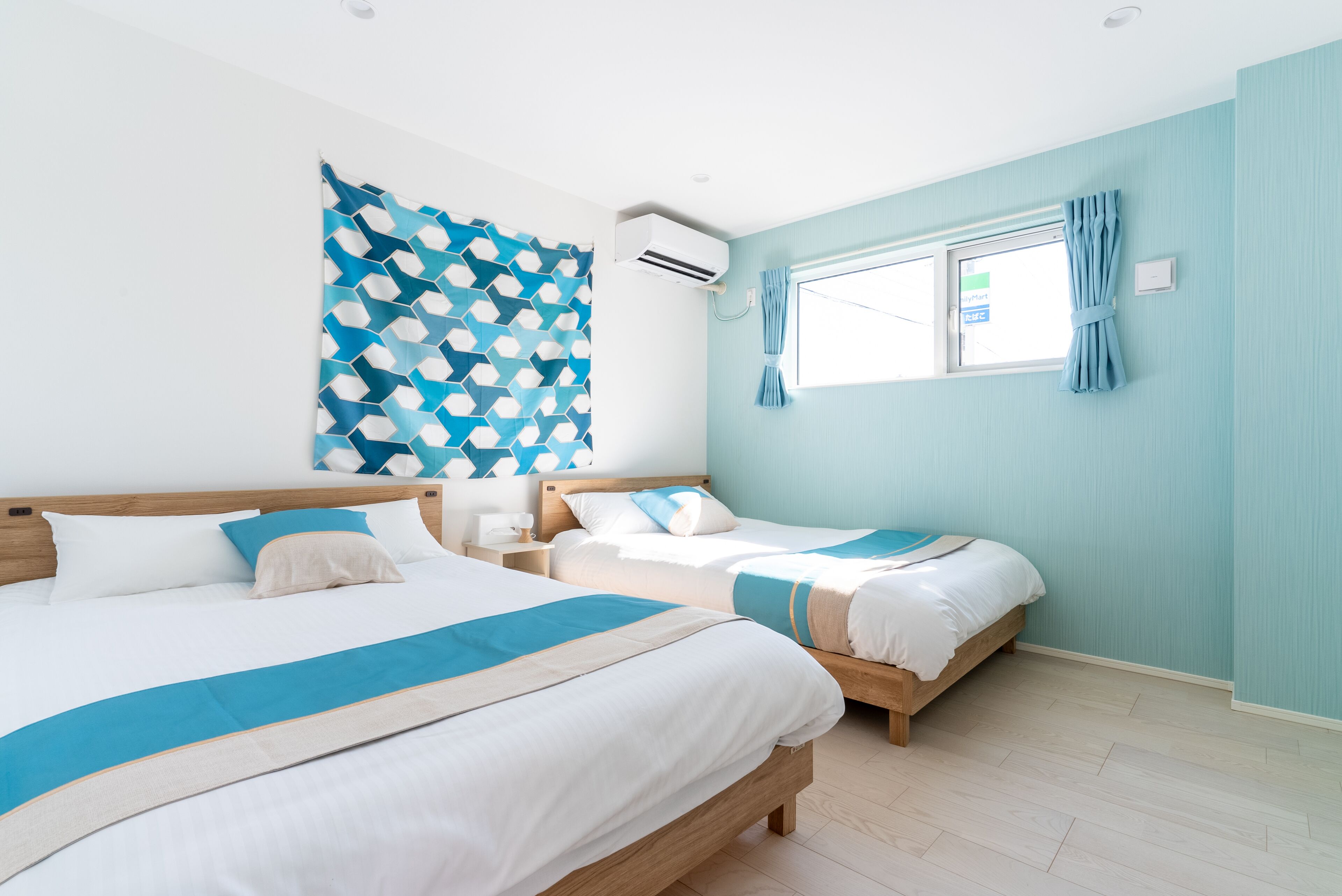 A hotel room with two beds, a large window with curtains, an air conditioning unit, and a geometric-patterned wall hanging.