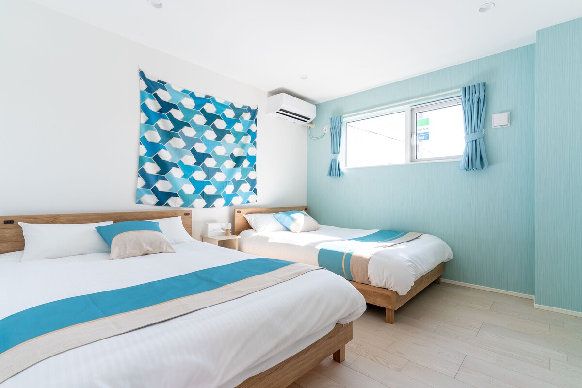 A hotel room with two beds, a large window with curtains, an air conditioning unit, and a geometric-patterned wall hanging.