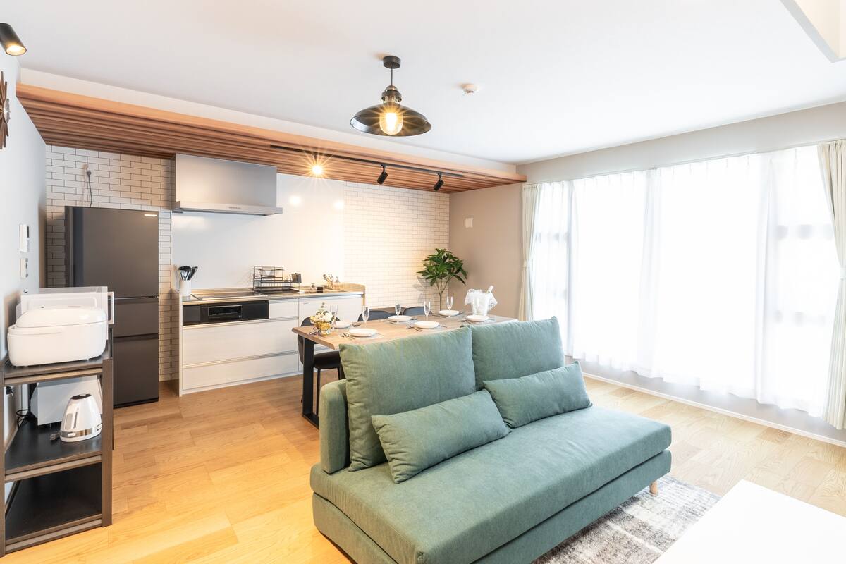 A modern kitchen with a built-in fridge, a dining area with a table and chairs, and a green sofa in a bright living room with large windows.