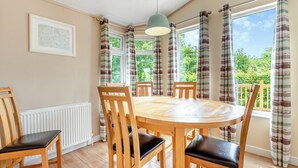 A dining room with a round table, six wooden chairs, a hanging light fixture, and large windows with curtains. - Stylish luxury lodge with meadow view. (Wattleborough, Nr Shrewsbury)