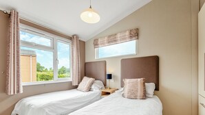 A bedroom with two single beds, a nightstand with a lamp, a window with curtains, and a view of greenery outside. - Stylish luxury lodge with meadow view. (Wattleborough, Nr Shrewsbury)