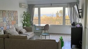 Uma sala de estar com um sofá bege, uma mesa de jantar com cadeiras, uma janela grande com cortinas e uma planta.