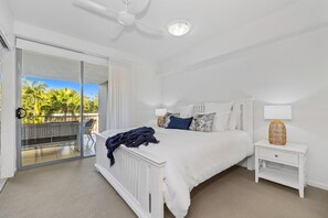 Relax in a serene bedroom with a large bed, nightstand, and ceiling fan. Enjoy a balcony with a view of palm trees.