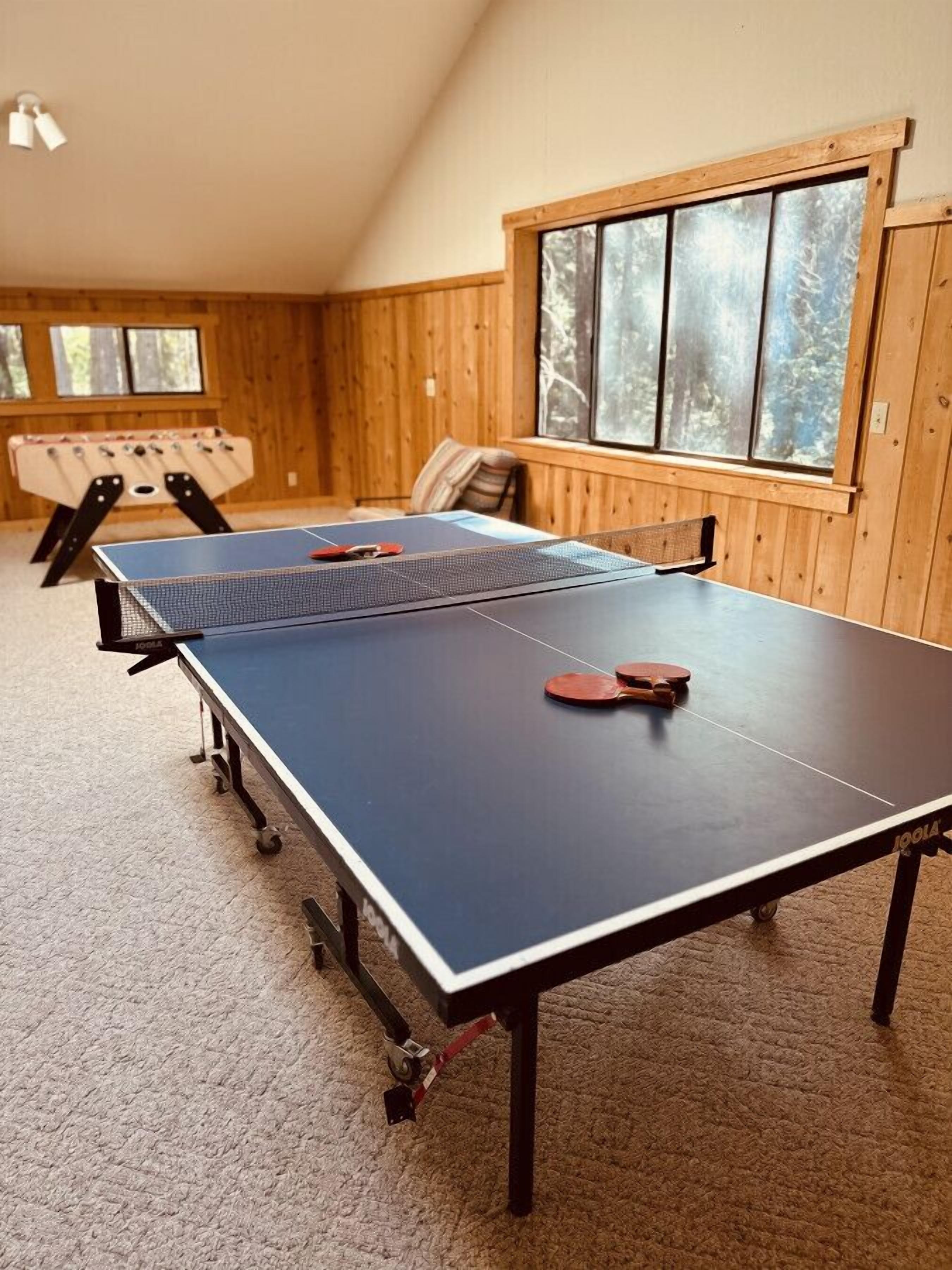 A ping pong table with two paddles, a foosball table, and a window with a view of trees.