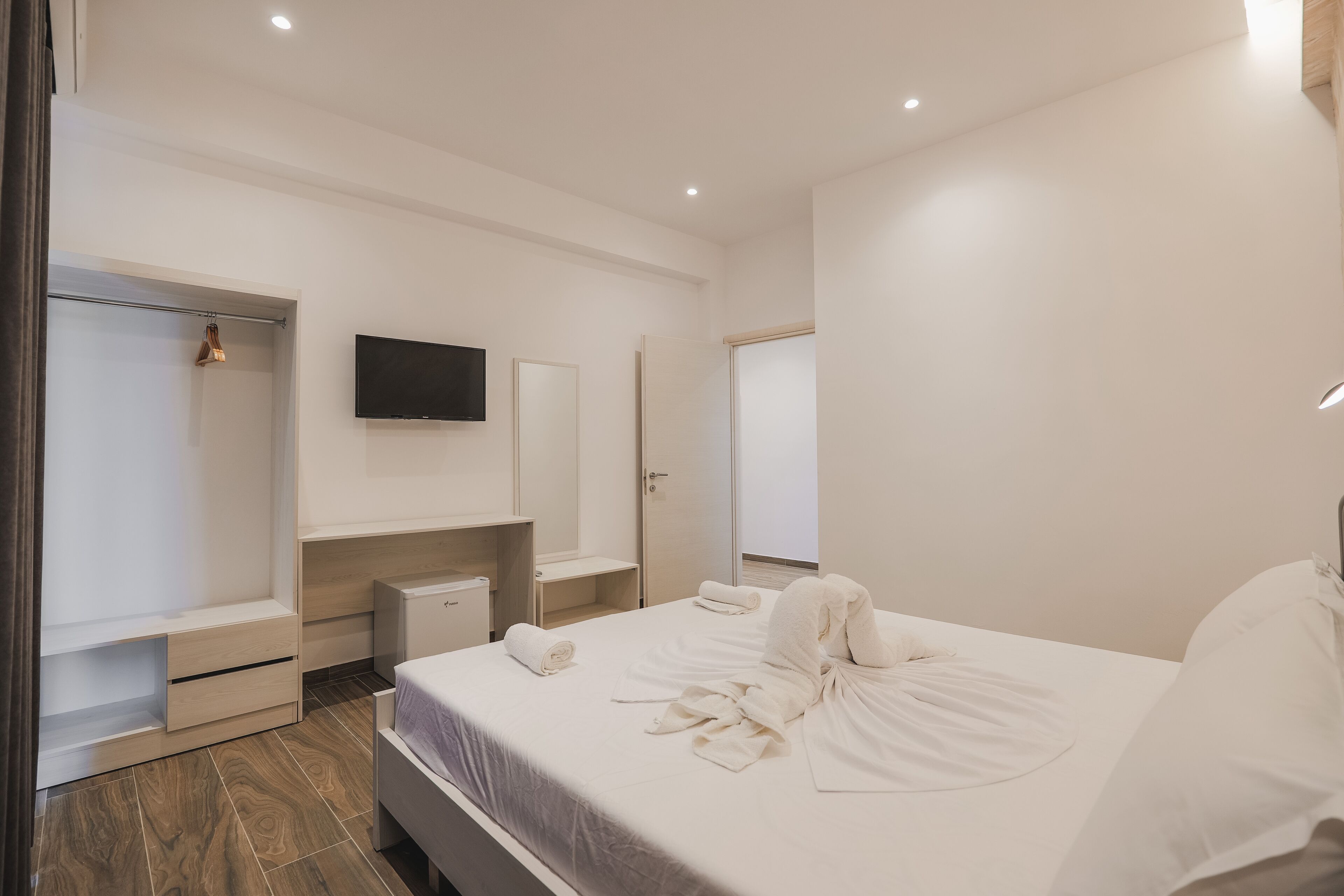 A modern hotel room with a large bed, a flat-screen TV, a wardrobe, and a small bedside table.