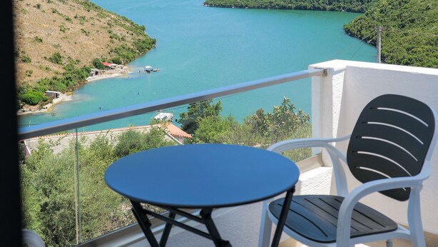Un balcón con mesa y sillas con vistas a un lago turquesa y colinas verdes.