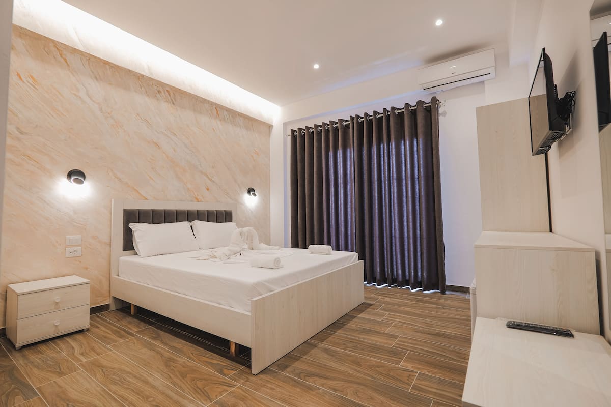 Quarto moderno com cama grande, mesas de cabeceira, televisão na parede e janela com cortinas.
