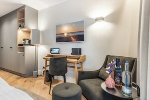 Quarto de hotel moderno com escrivaninha de madeira, computador, televisão, sofá e uma pequena mesa com garrafas.