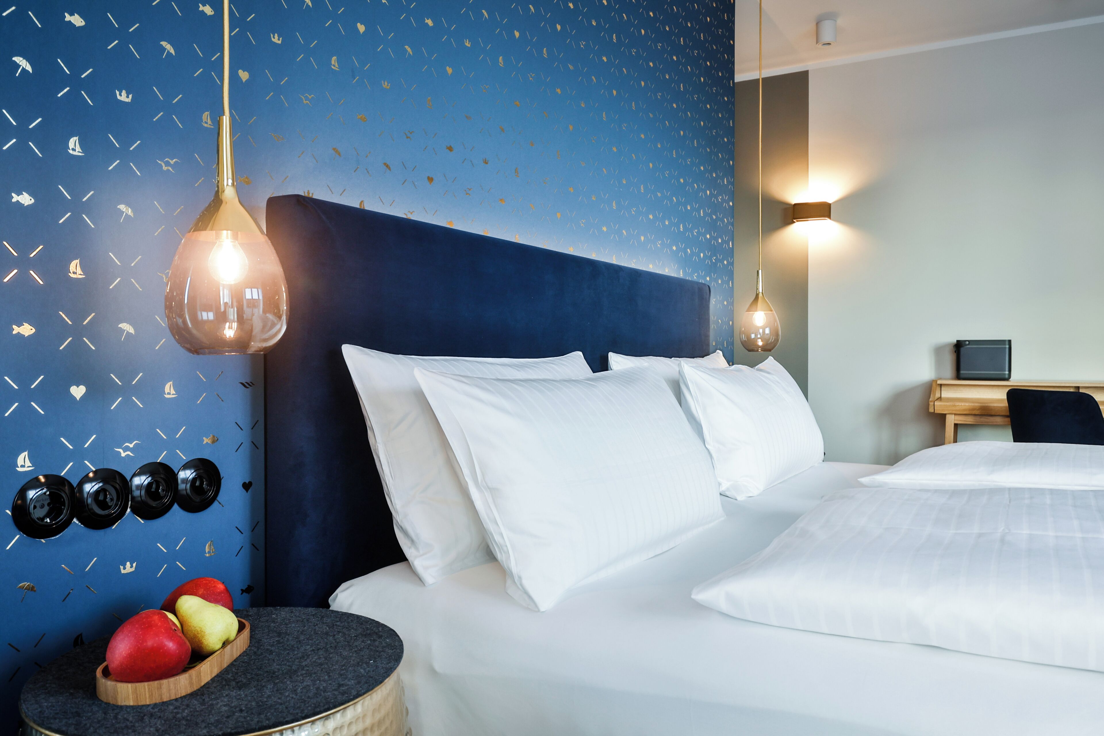 Una cama bien tendida con sábanas blancas, pared azul con papel tapiz de temática náutica y una mesita de noche de madera con manzanas.