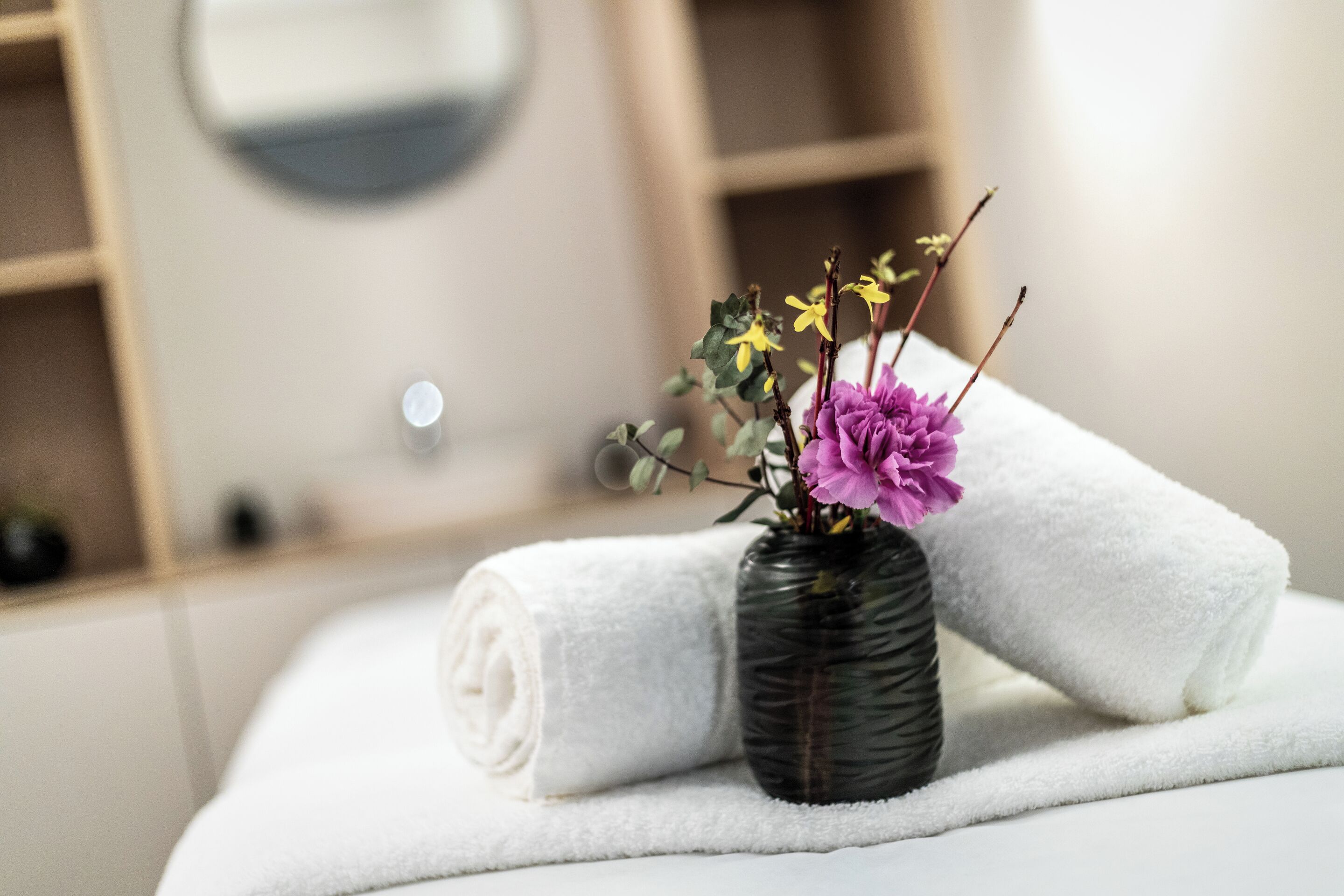 A neatly folded white towel on a flat surface, a dark glass vase with purple and yellow flowers, and a blurred background suggesting a bathroom setting.