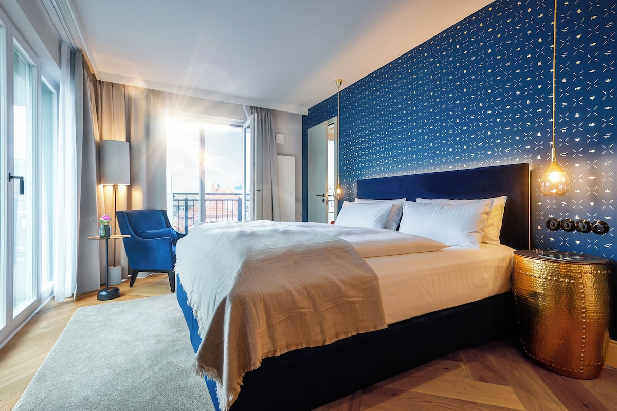 A modern hotel room with a large bed, a blue chair, a round side table, and a view of the city through large windows.