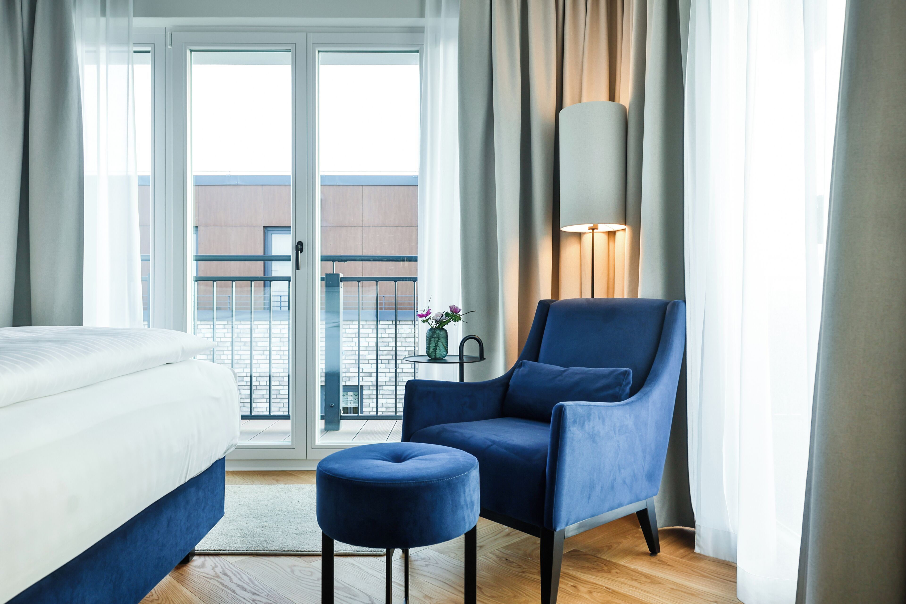 Un cuarto de hotel con cama, un sillón azul, un pequeño reposapiés, una lámpara y vistas de un edificio a través de la puerta de vidrio.