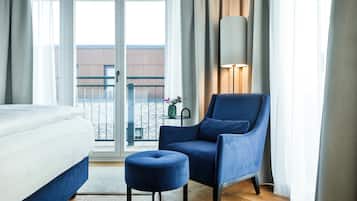 Et hotelværelse med en seng, en blå lænestol, en lille skammel, en lampe og udsigt til en bygning gennem glasdøren.