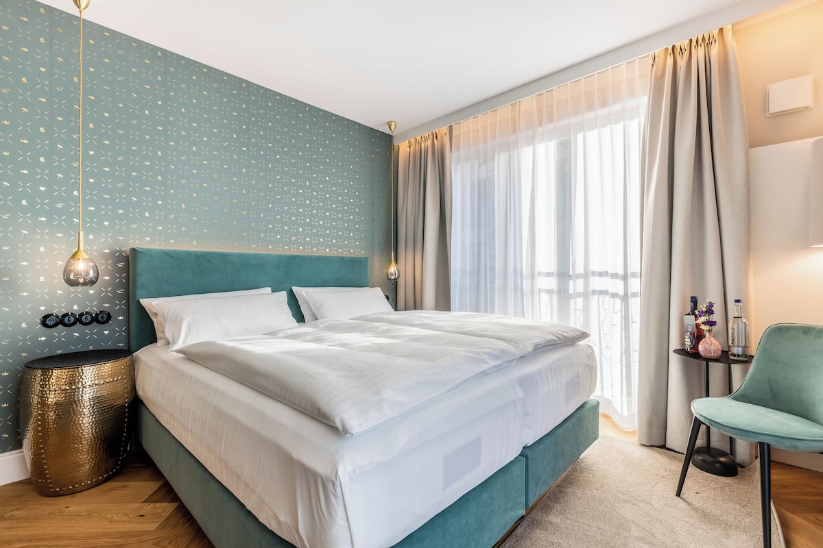 A neatly made bed with white linens, a teal headboard, and a small bedside table with a vase and bottles.