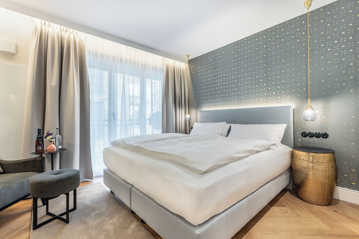 A modern bedroom with a large bed, a side table, a small stool, and a wall-mounted light fixture.