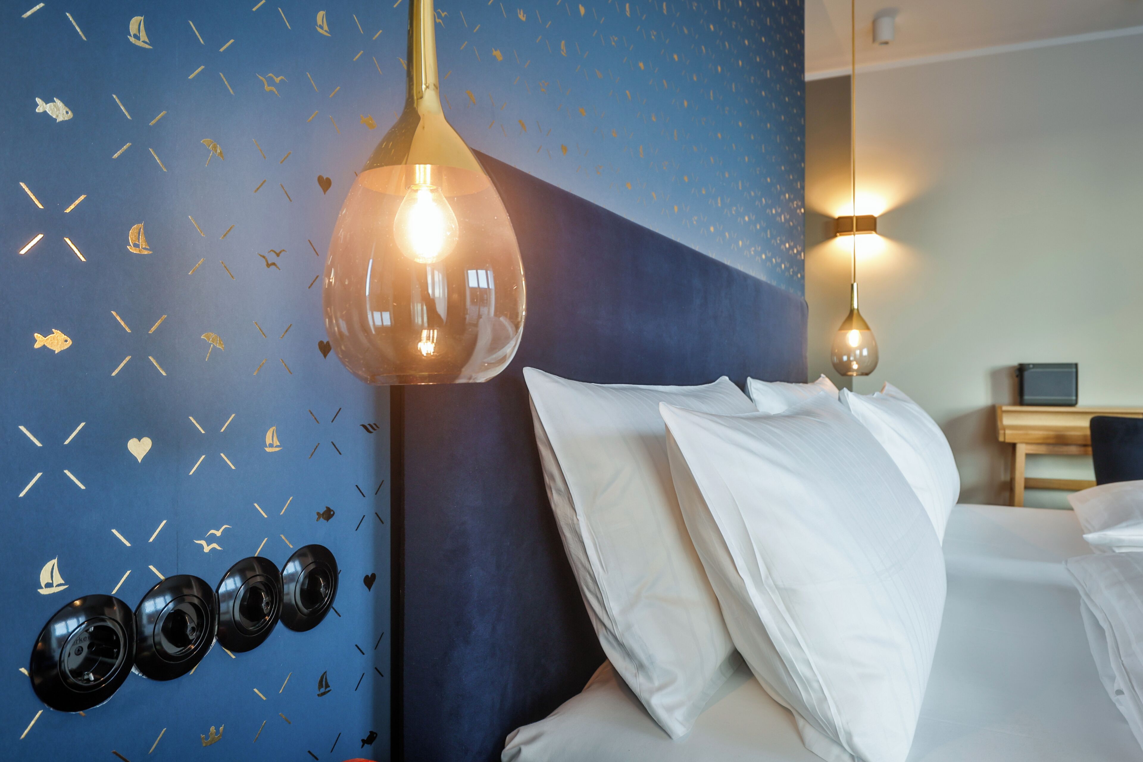 Habitación de hotel con pared azul decorada con motivos náuticos, cama con ropa blanca y lámpara colgante.