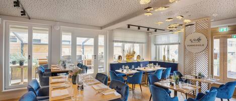 Ein modernes Restaurantambiente mit blauen Samtstühlen, Holztischen und einer Decke mit strukturierten Lampen.