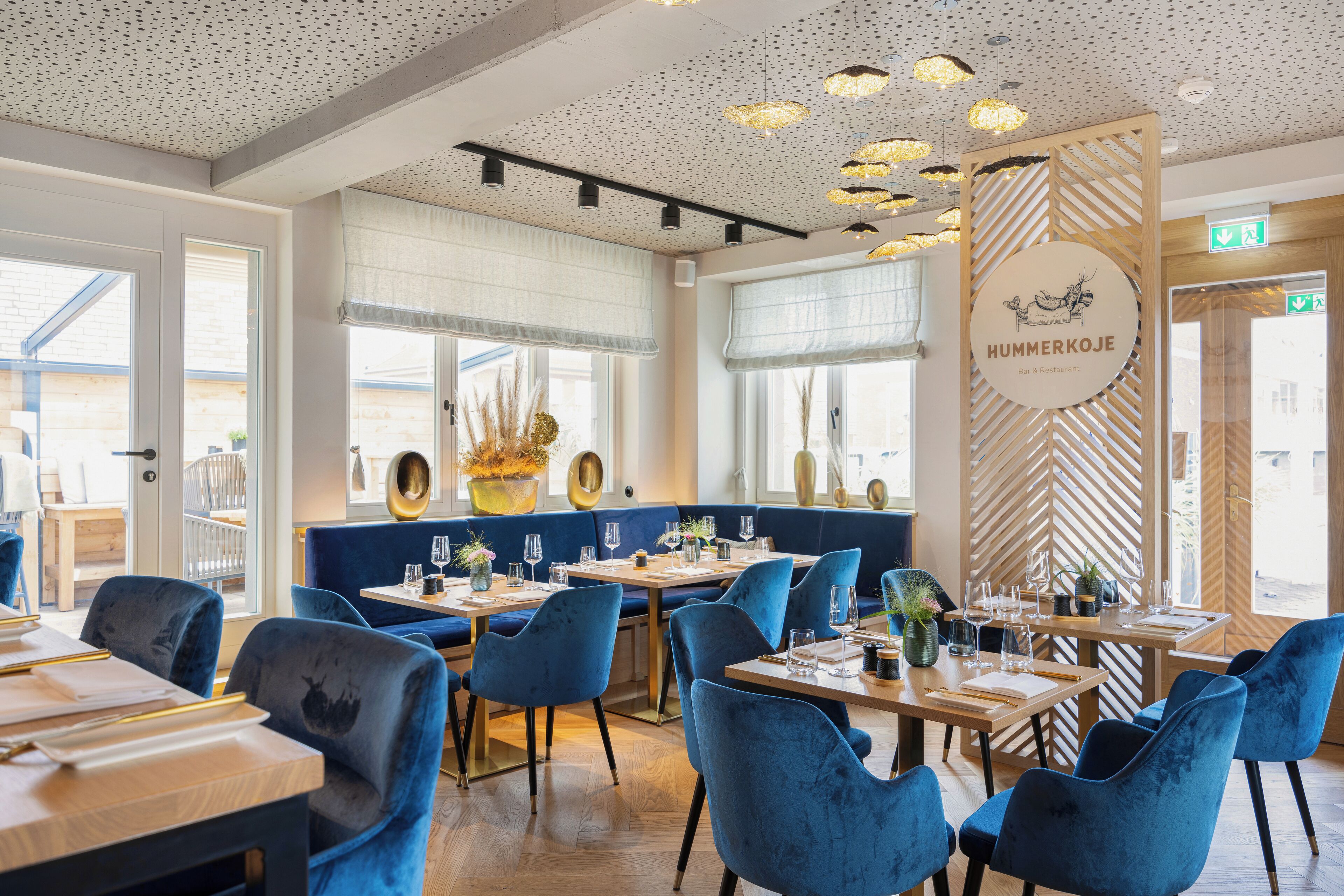 A restaurant interior with blue velvet chairs, wooden tables, and a herringbone patterned wall.