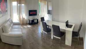 A modern living room with a white sofa, a dining table with four chairs, and a TV displaying the Netflix logo.