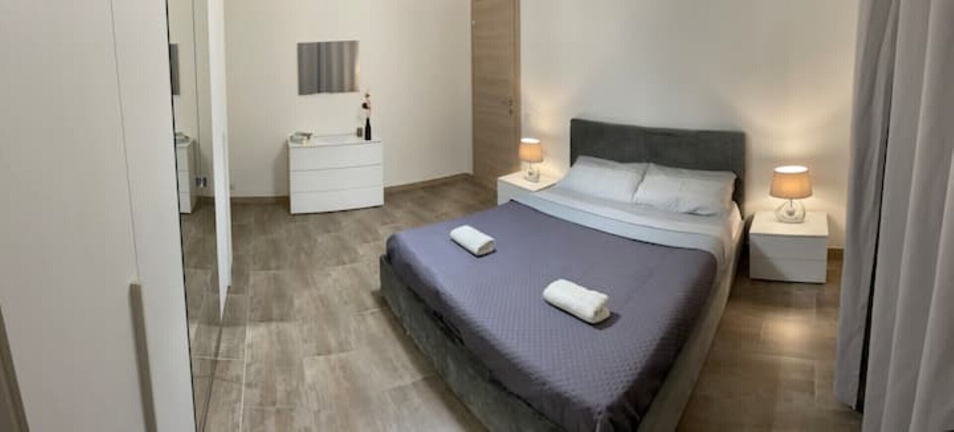 Un dormitorio con cama, dos mesitas de noche, un tocador y una lámpara de pared.