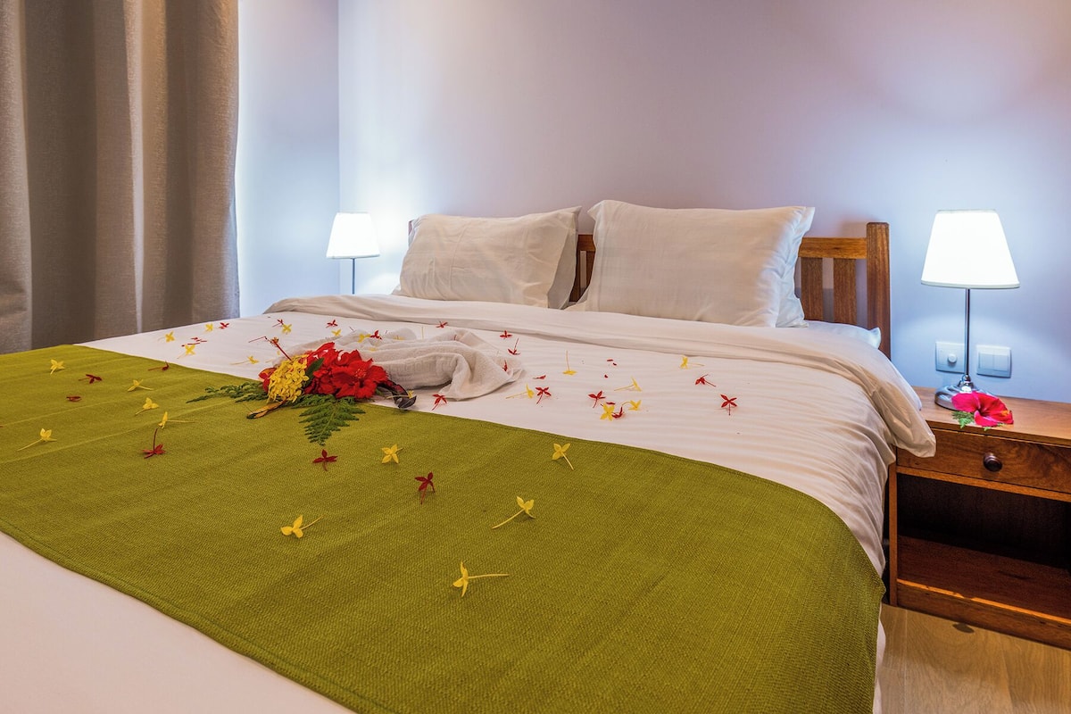Una cama bien hecha con una colcha verde, almohadas blancas y un arreglo floral. Hay dos mesitas de noche con lámparas.