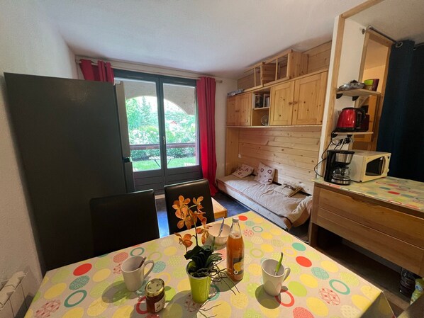 A compact room with a dining table, a sofa, a kitchenette, and a window with a view of greenery.