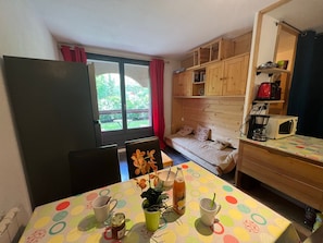 A compact room with a dining table, a sofa, a kitchenette, and a window with a view of greenery.