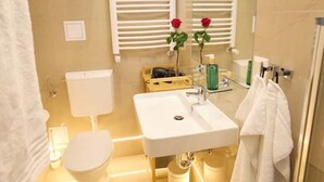 Ein modernes Badezimmer mit einem weißen Waschbecken, einer Toilette und einem Bidet. Es gibt zwei weiße Handtuchhalter, einen Spiegel und eine rote Rose am Waschbecken.