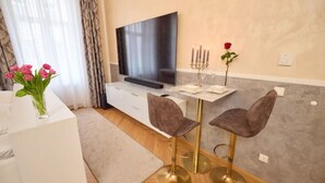 Ein modernes Wohnzimmer mit Flachbildfernseher, einem weißen Sofa, einem Couchtisch, zwei Barhocker, einer Blumenvase und einem dekorativen Kerzenleuchter.
