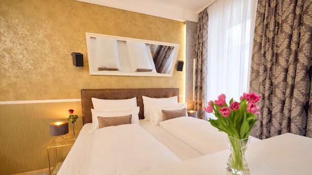 Ein Hotelzimmer mit einem großen Bett, einem Nachttisch mit Lampe, einer Blumenvase und einem Fenster mit Vorhängen.