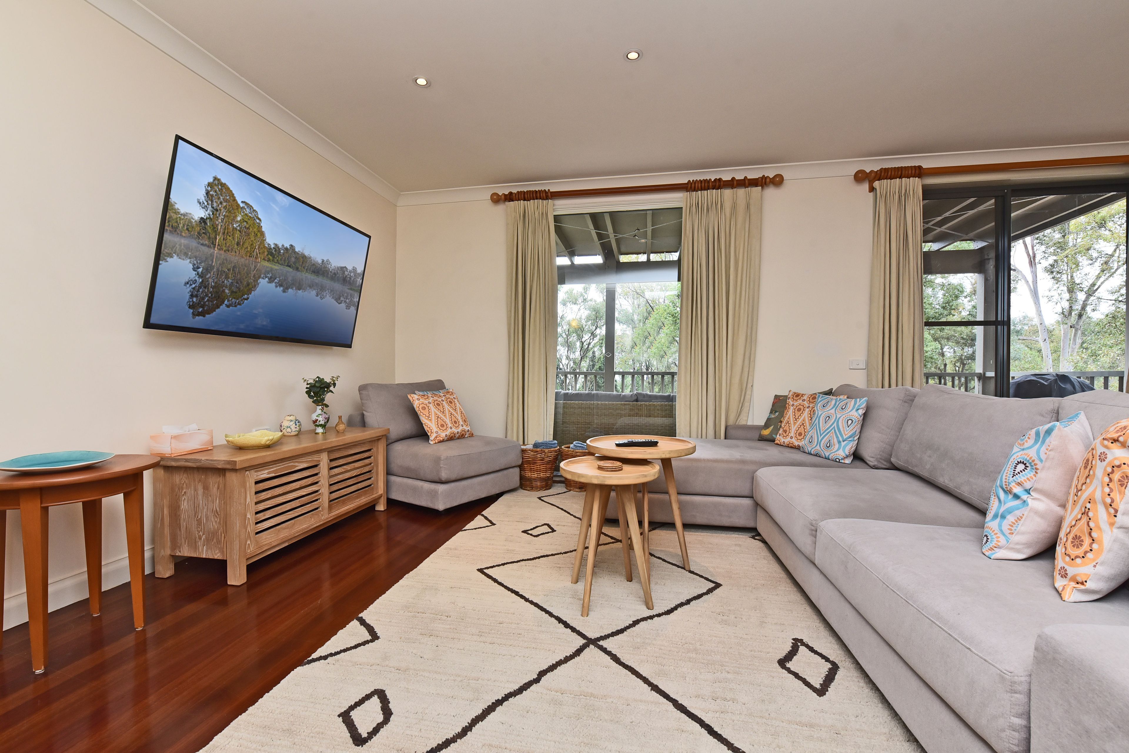 A living room with a grey sofa, a wooden coffee table, a TV mounted on the wall, and a large window with curtains.