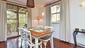 Un comedor con mesa de madera, sillas blancas, lámpara colgante y vista a una terraza exterior a través de una puerta de cristal.