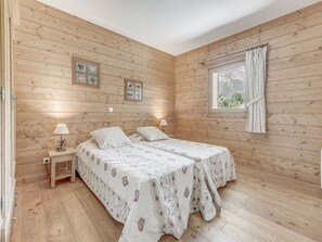 Une pièce aux murs en bois, avec deux lits dotés de draps à motifs, une table de chevet avec une lampe et une fenêtre avec des rideaux.