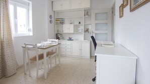 A small, bright room with white walls and a window. There is a white desk with a chair, a white cabinet with shelves, and a small table with a chair. - RentalSevilla in the Triana neighbourhood (Sevilla)