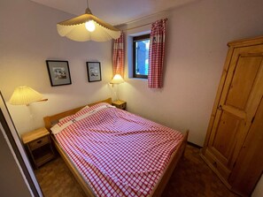A bedroom with a bed, two bedside lamps, a window with curtains, a wooden wardrobe, and two framed pictures on the wall.