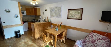 A compact kitchen with wooden cabinets, a microwave, and a coffee maker. There is a dining table with chairs and a small potted plant. A television is mounted on the wall.