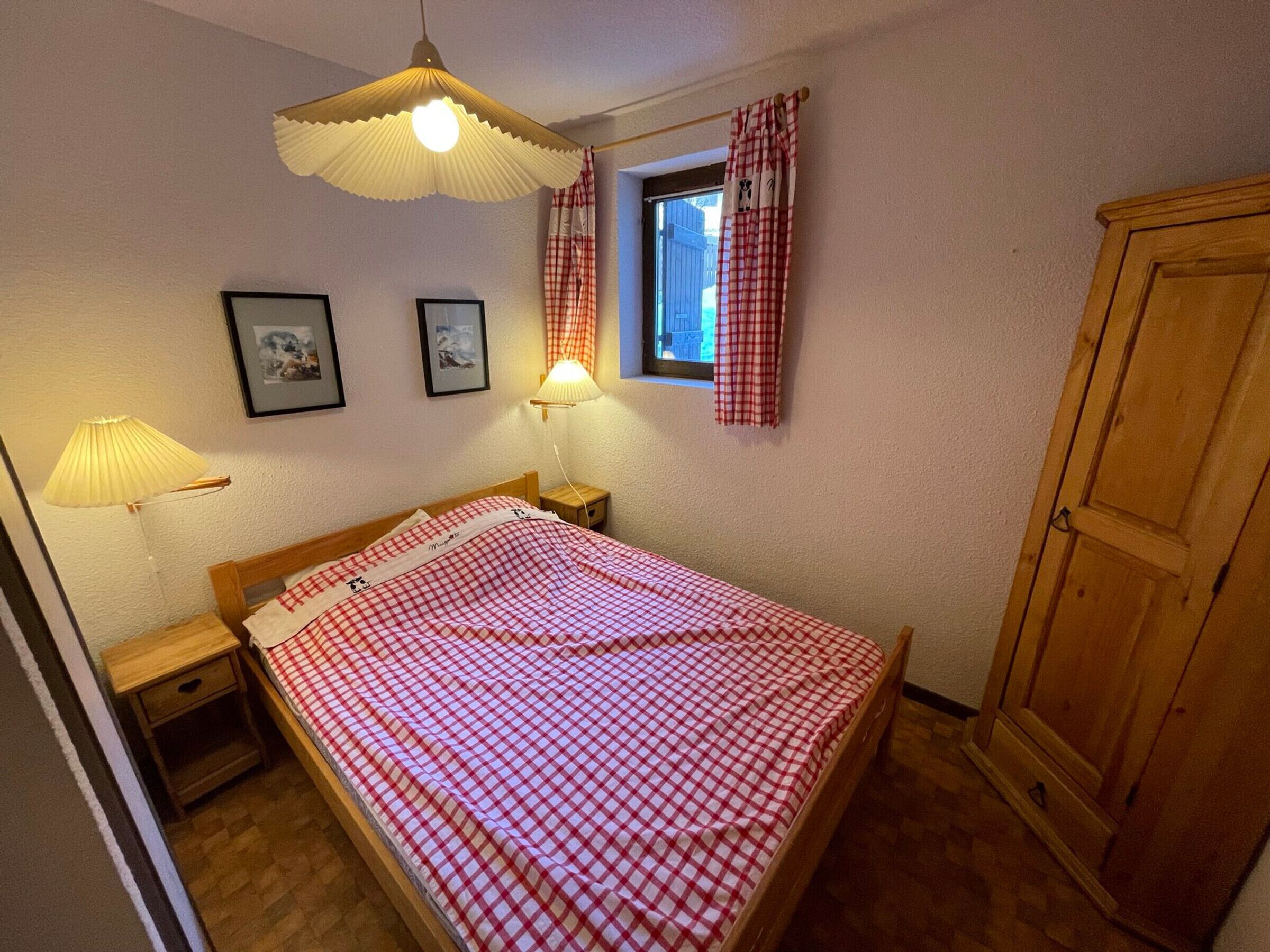Een slaapkamer met een bed, twee bedlampen, een raam met gordijnen, een houten garderobe en twee ingelijste foto's aan de muur.