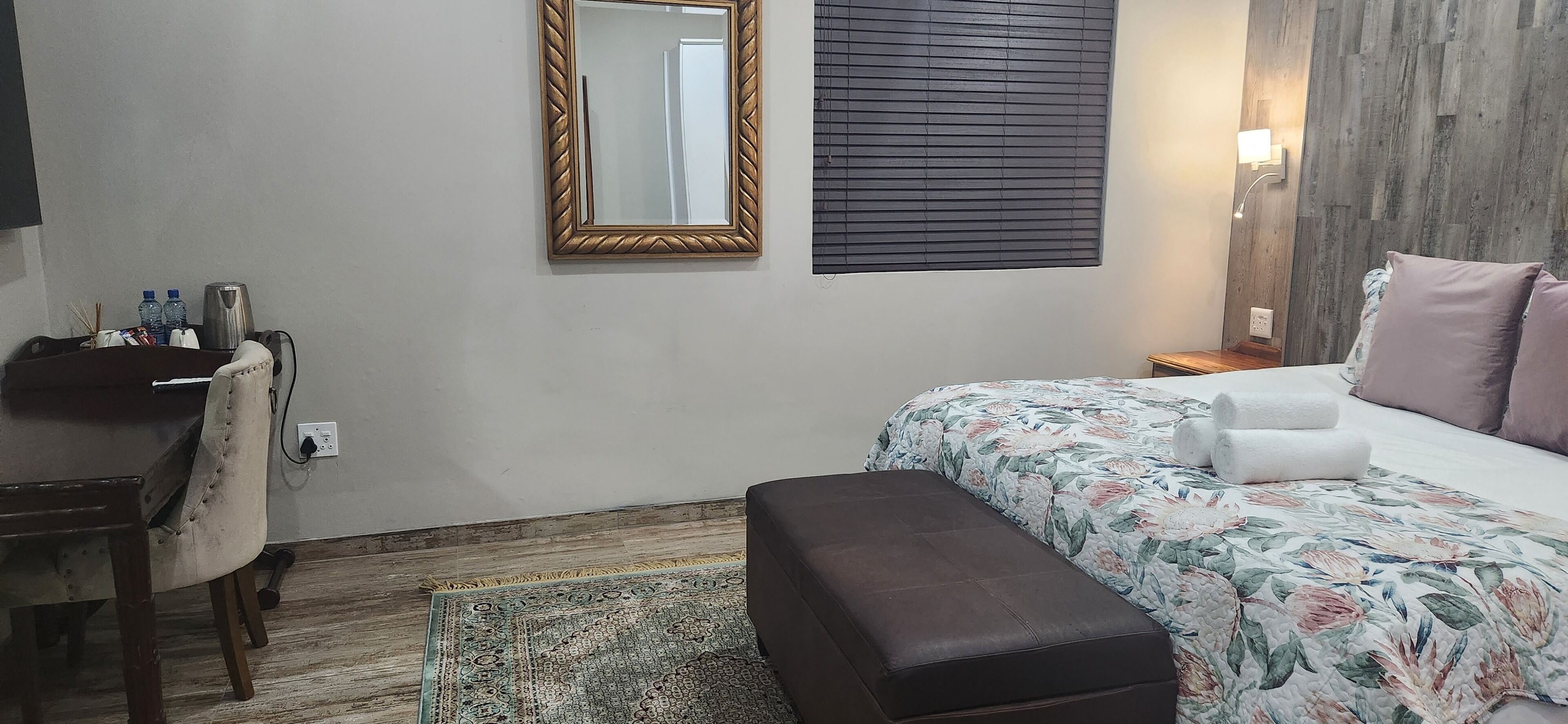 A bedroom with a bed, a bench, a desk, a mirror, and a window with blinds.