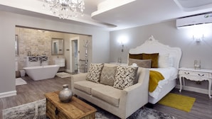Un dormitorio moderno con una cama grande, una zona de estar con sofá, una mesa de centro de madera y un baño a la vista al fondo.