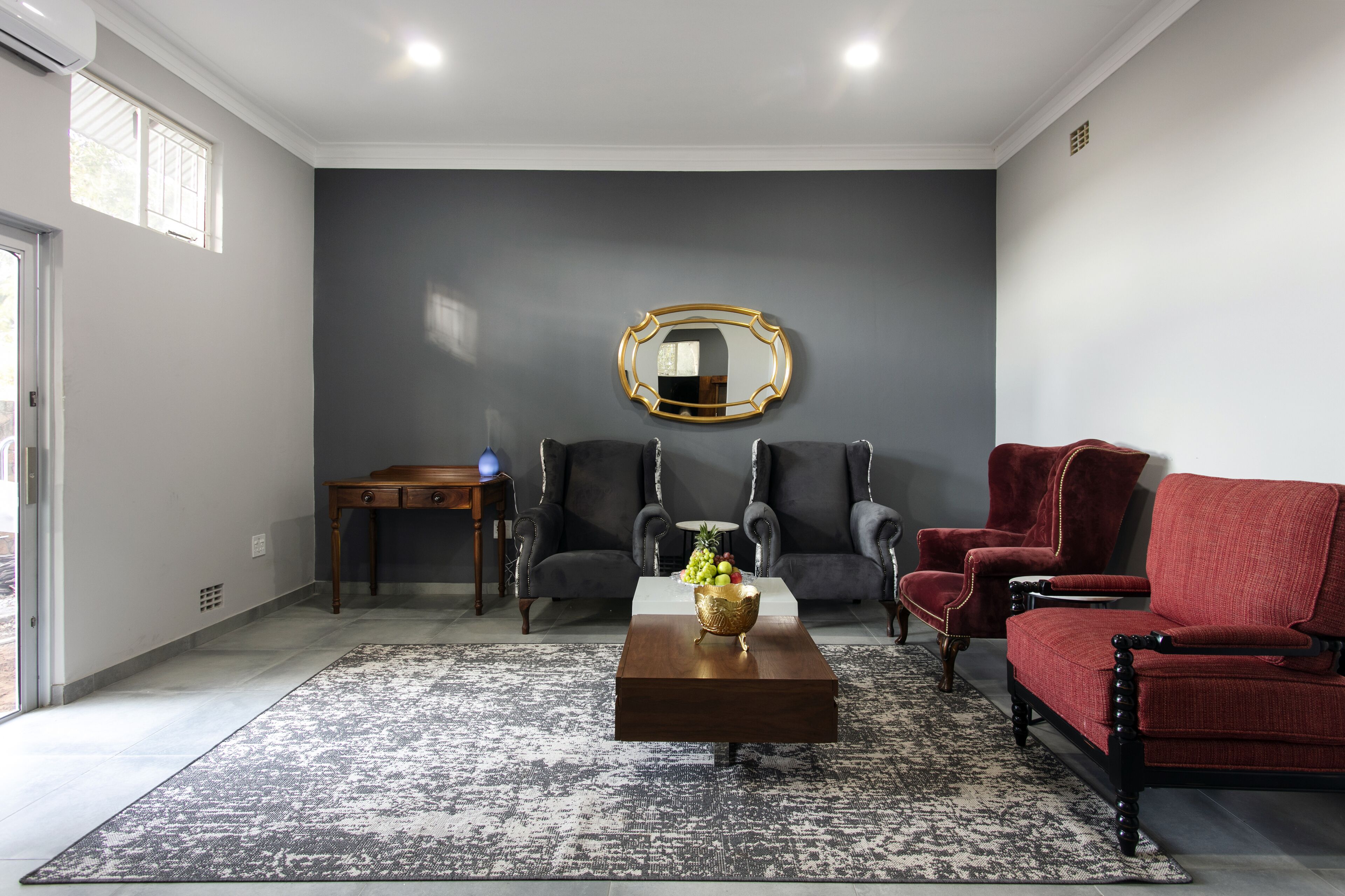 A living room with a dark gray wall, a round mirror, two armchairs, a red armchair, a wooden coffee table, and a patterned rug.
