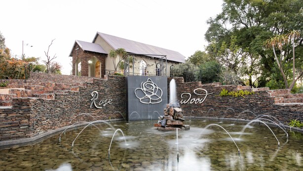 Una fuente de piedra con chorros de agua en una piscina cristalina, rodeada por un muro de piedra con un diseño de cruz y rosa, y un edificio con techo inclinado al fondo.