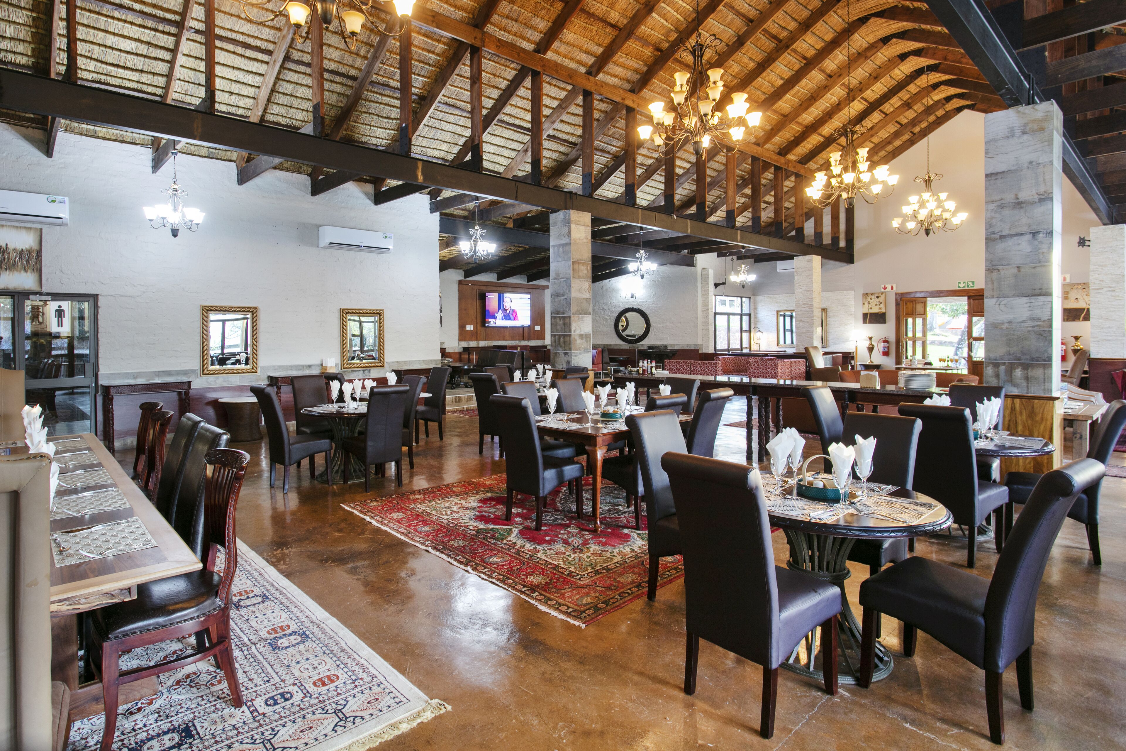 A spacious restaurant with high ceilings, wooden beams, and chandeliers. Tables are set with chairs and centerpieces.