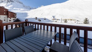 Un balcon doté d’une balustrade en bois, d’une table et de chaises, offrant une vue sur un paysage montagneux enneigé, avec des bâtiments et des remontées mécaniques.