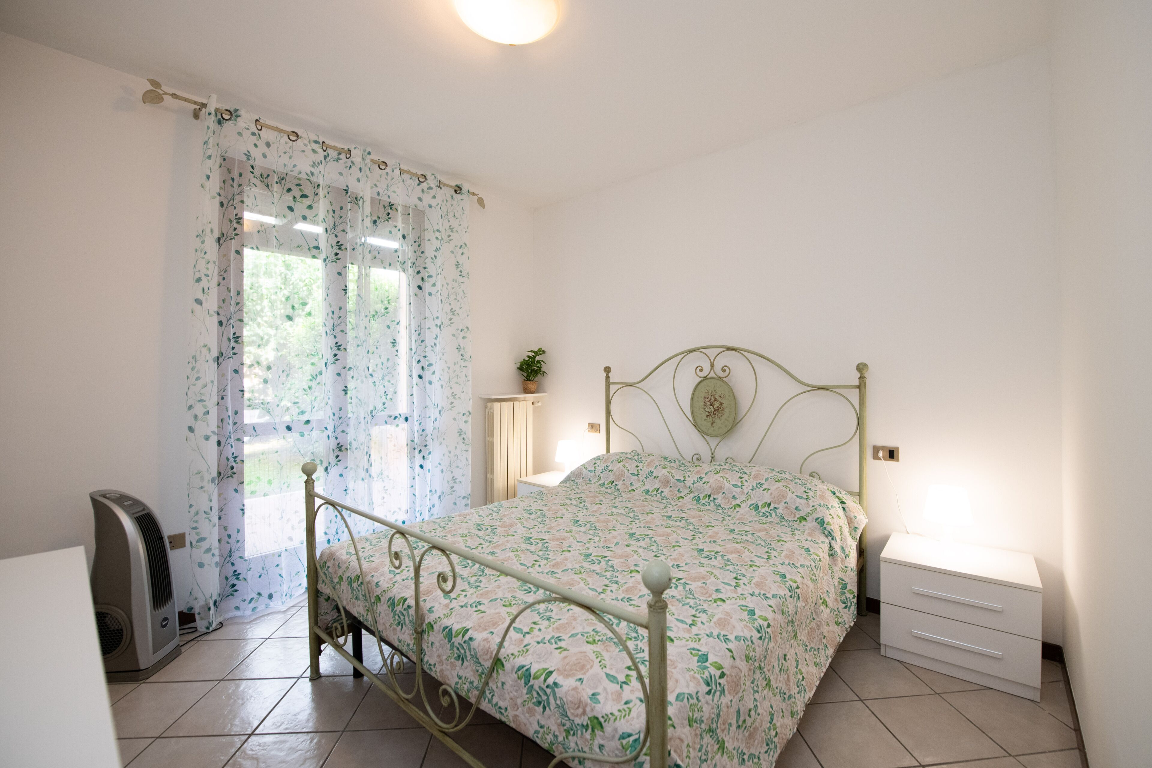 Una camera da letto con una struttura letto in metallo, un comodino con cassetti, un termosifone e una finestra con tende floreali.