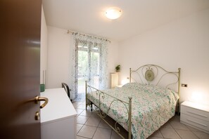 Um quarto com estrutura de cama em metal, mesa de cabeceira, uma cadeira e uma escrivaninha.