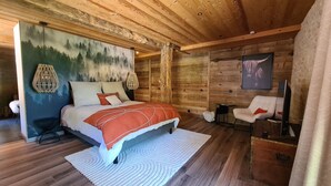 Camera da letto accogliente di una baita di legno, con un letto, una sedia e un quadro raffigurante una foresta.
