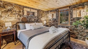 A cozy bedroom with a stone wall, a wooden ceiling, and a bed with a grey blanket. - Historic | Int Designed | DT Flag | Atrium | AC (Flagstaff)